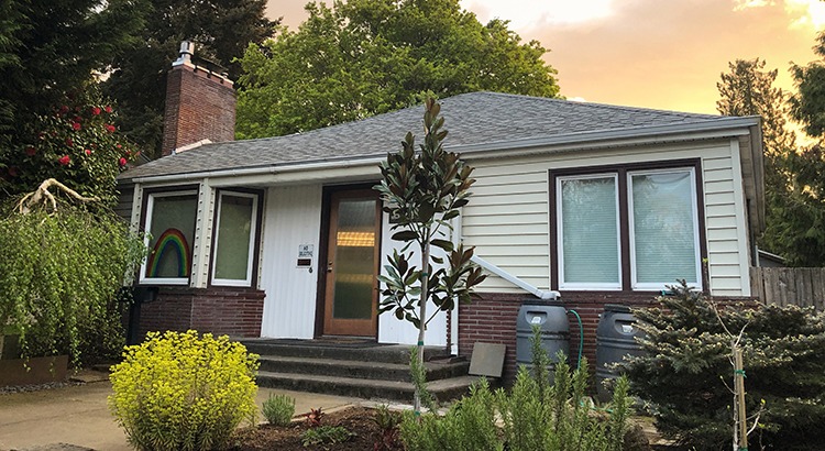 3/4 on shot of a 1940’s mid century bungalow style home with a dramatic sunset behind it and a rainbow painted in the window. Portland, OR